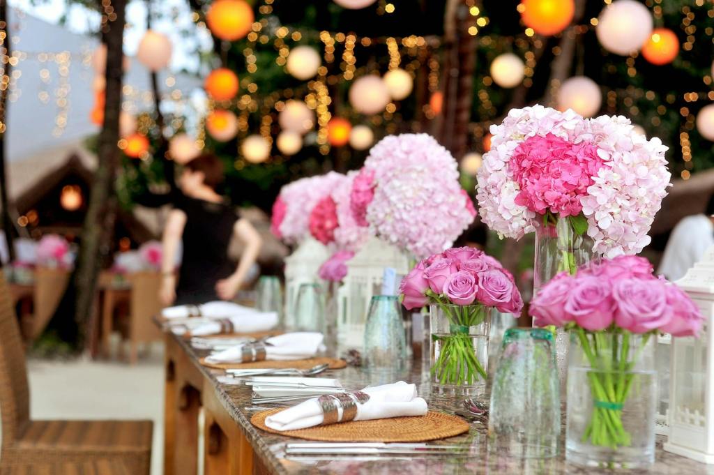 A table set with cutlery, napkins and glasses and brightly decorated with flowers in a variety of shades of pink. In the background, lights and decorations are hung up.