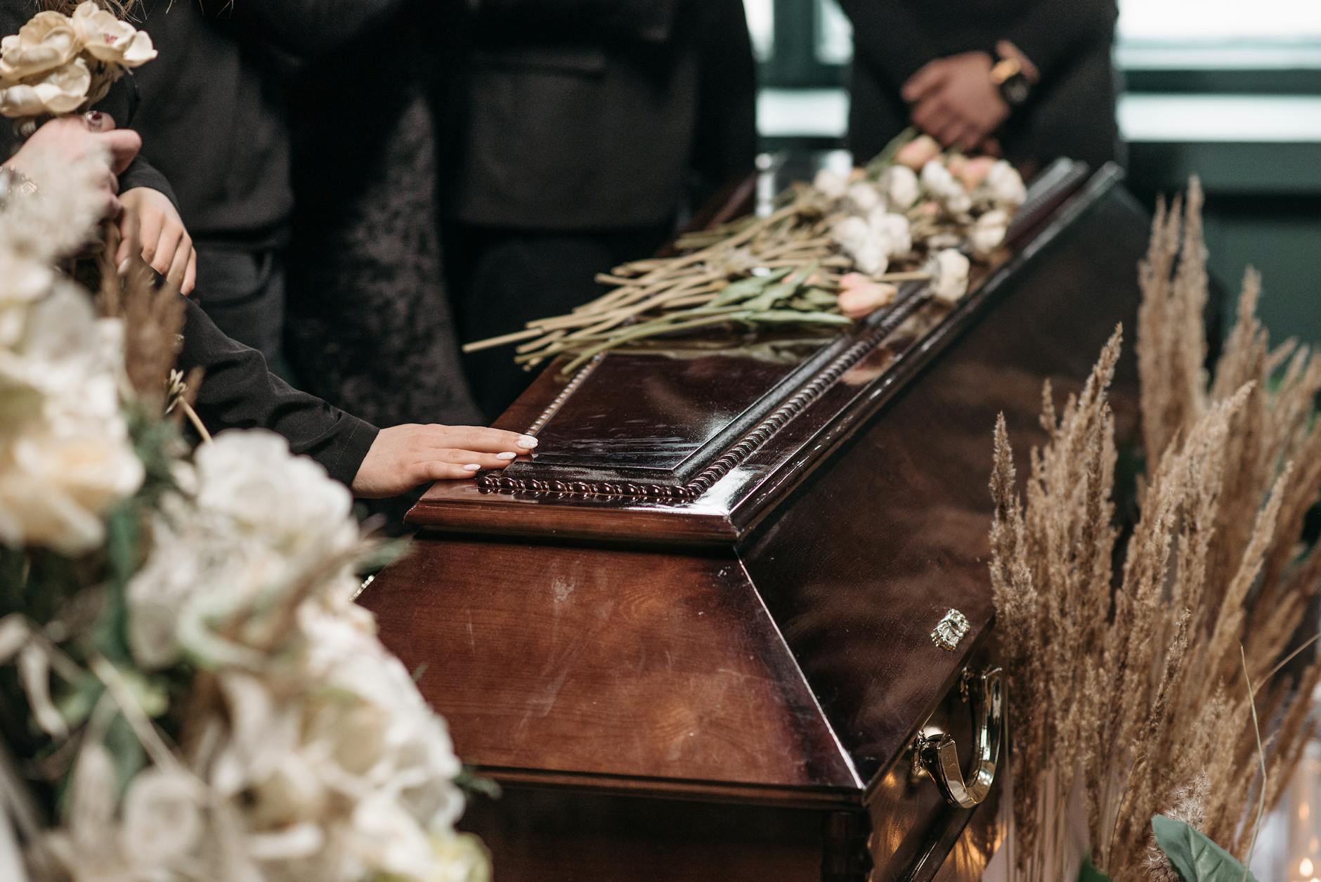 A brown wooden coffin runs diagonally from bottom left to top right, with flowers to the left, right and on top of the coffin. The hands of several mourners are visible including one touching the top of the coffin.